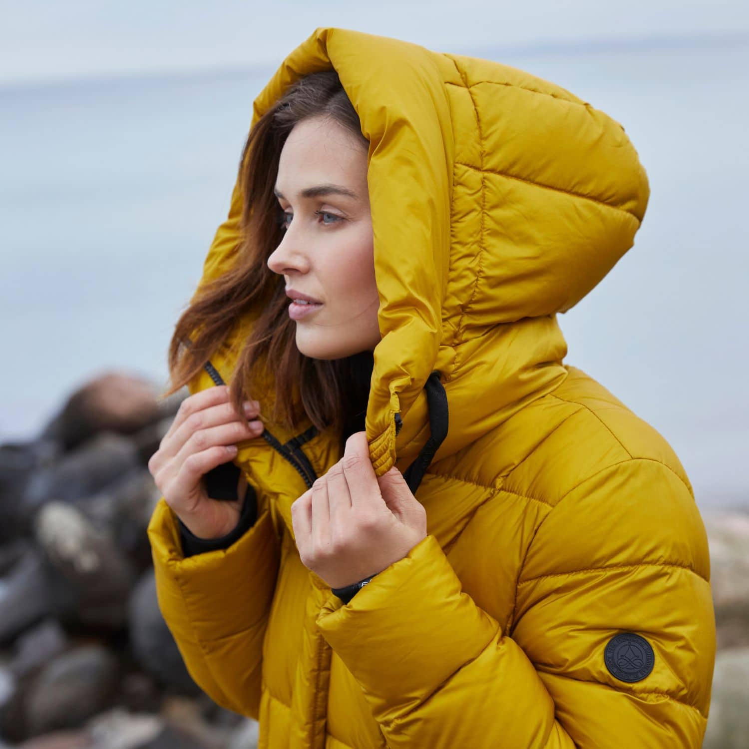Model i gul efterårsjakke med hætte ved havet, der viser efterårsfarver for 2025 og tidens jakke trends